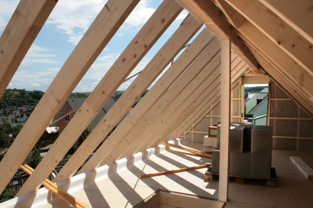 Construction Charpente Bois : Aménagement de Combles Charpente en bois d'un grenier en construction, vue intérieure avec lumière extérieure et aperçu d'un village vallonné.