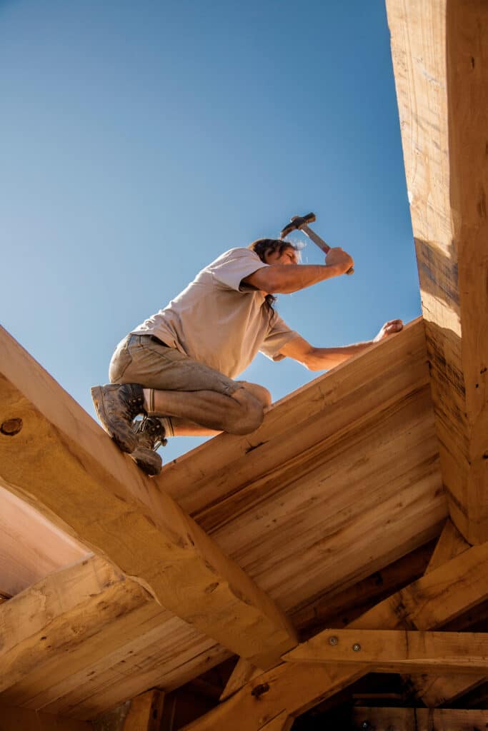 Charpentier au travail : construction ossature bois sous ciel bleu Charpentier accroupi sur une poutre en bois, martelant sous un ciel bleu clair, construction de charpente.