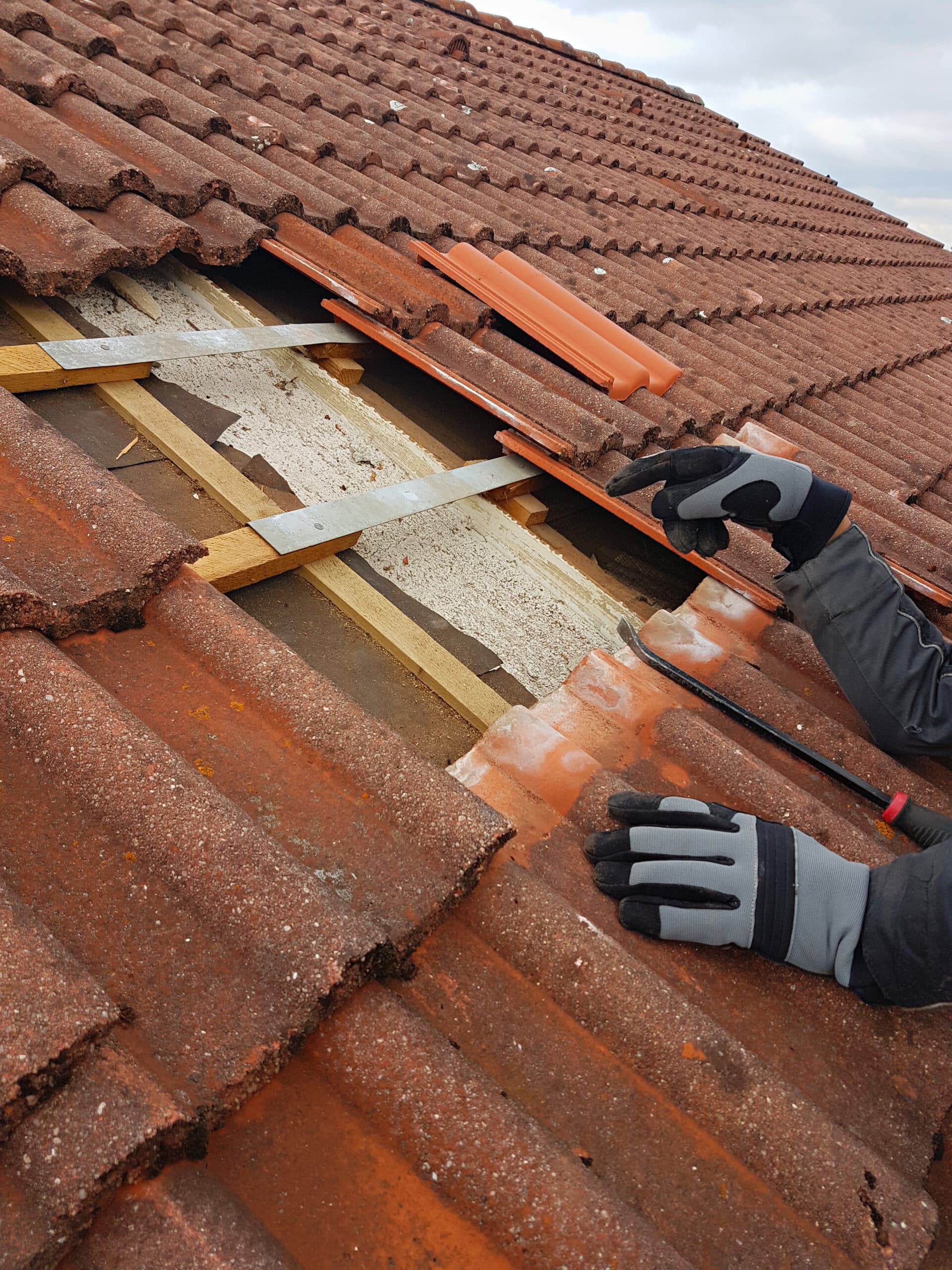 Réparation Toiture : Enlèvement Tuiles et Accès Sous-face Un ouvrier avec des gants gris et noirs enlève une tuile de toit en terre cuite rouge et granuleuse pour accéder à la sous-toiture et aux liteaux.