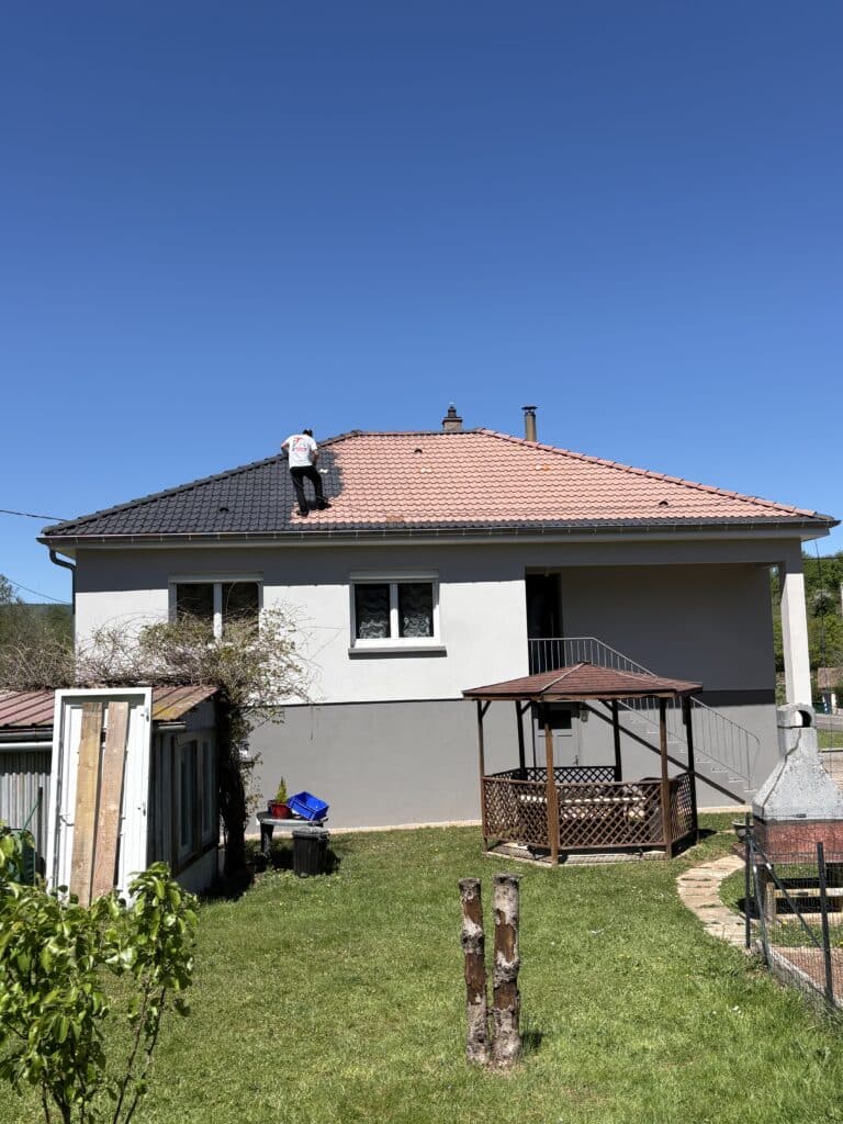 Peinture de toiture : Rénovation de tuiles en gris Artisan peignant le toit d'une maison individuelle en gris anthracite sous un ciel bleu ensoleillé.